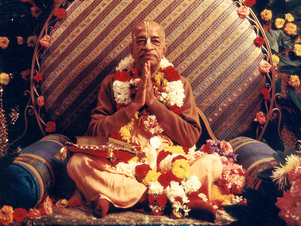 His Divine Grace A.C. Bhaktivedanta Swami Prabhupada in traditional saffron robes, sitting in meditation pose with serene expression and warm lighting